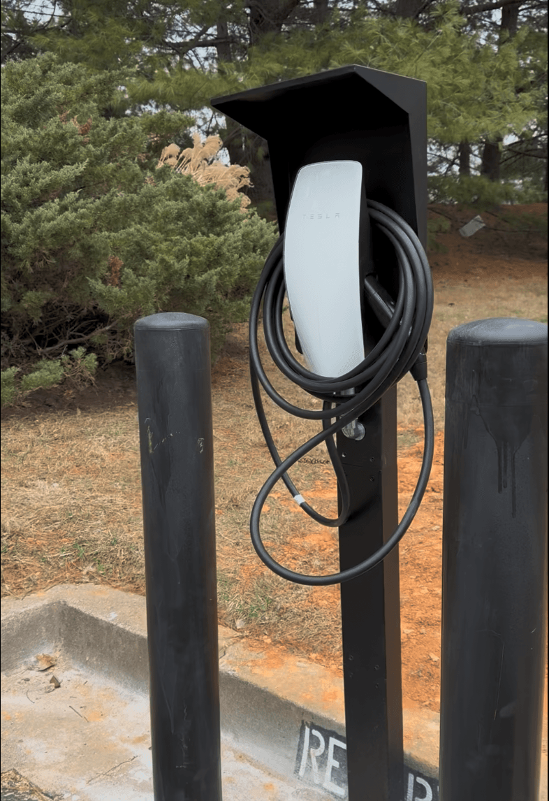 White Tesla wall charger with a coiled black cable, mounted on a post between black bollards.